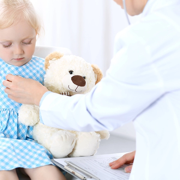 A child in a blue gingham dress holds a teddy bear while a healthcare professional examines them in a bright, clinical setting.