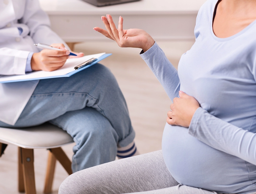 A pregnant woman sits and talks to a doctor, who is taking notes during a consultation in a medical office.